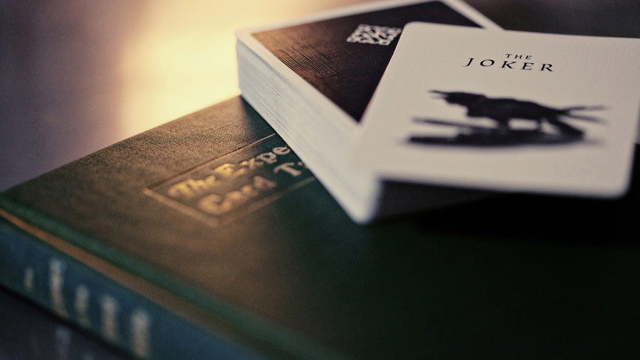 A close-up of Madison Black Rounders playing cards labeled 'THE JOKER' resting on top of a The Expert at the Card Table  book. 