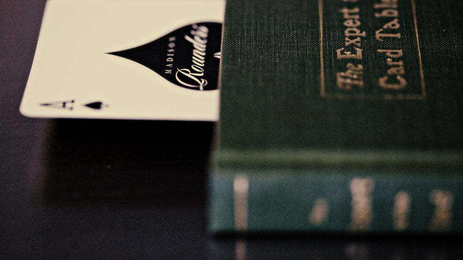 A close-up of a Madison Black Rounders  deck of cards with an Ace of Spades card partially tucked under a green hardcover book titled 'The Expert at the Card Table'.