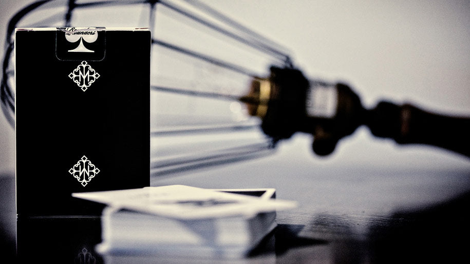 Madison Black Rounders tuck box with white spade and diamond logos stands on a reflective surface next to a stack of cards, with a blurred vintage-style card game apparatus in the background.
