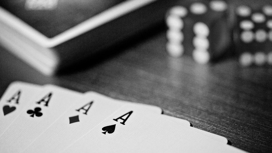 Four aces of a deck of Madison Black Rounders playing cards fanned out on a dark wooden table, with blurred dice in the background.