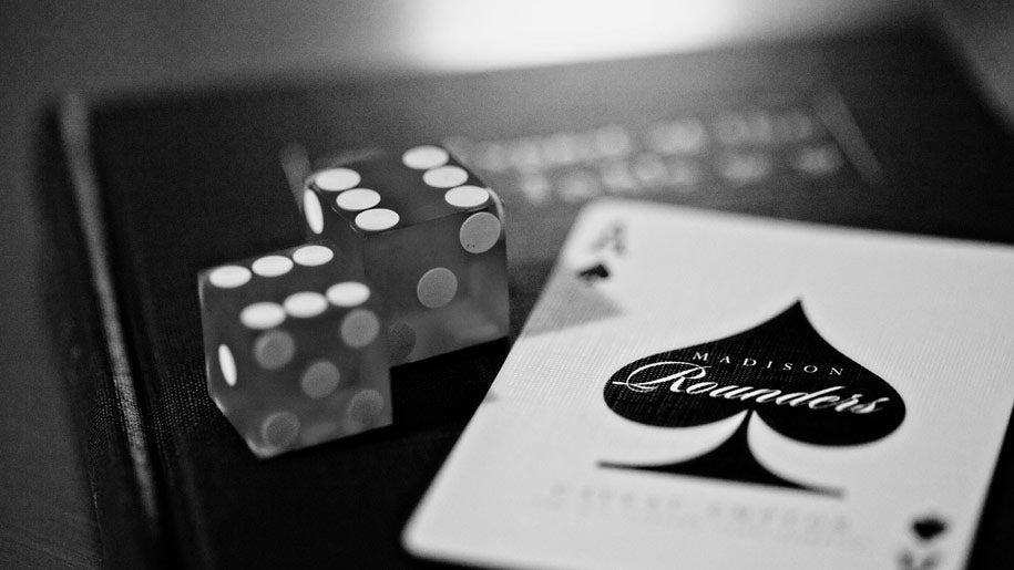 A black and white close-up of a Madison Black Rounders Ace of Spades, featuring two translucent dice showing white pips and an Ace of Spades card with the brand name 'Madison Readlers' printed on it, resting on a dark surface.